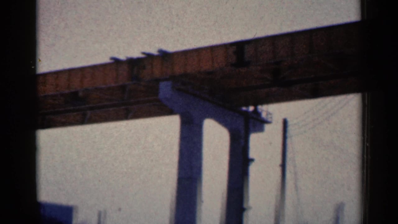1961 Duluth 8mm frame showing a dark railroad bridge span, vertical support, and faint utility lines above industrial surroundings.