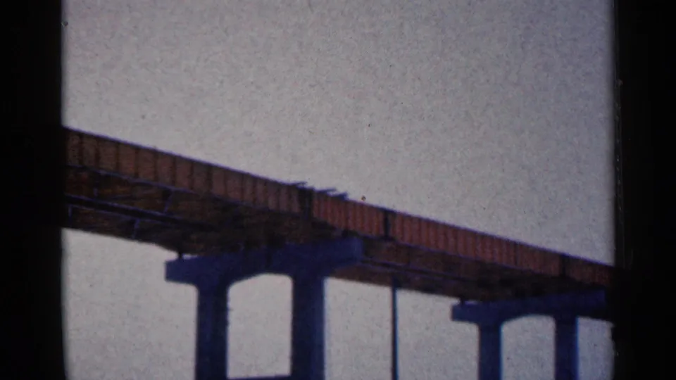 1961 Duluth frame showing the elevated brown bridge extending across the scene, with blue support columns, overhead wires, and pale overcast sky.