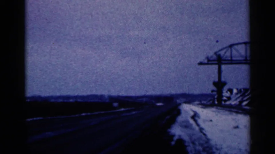 1961 Duluth travel frame showing a road or bridge approach with a structure at right and pale roadside ground.