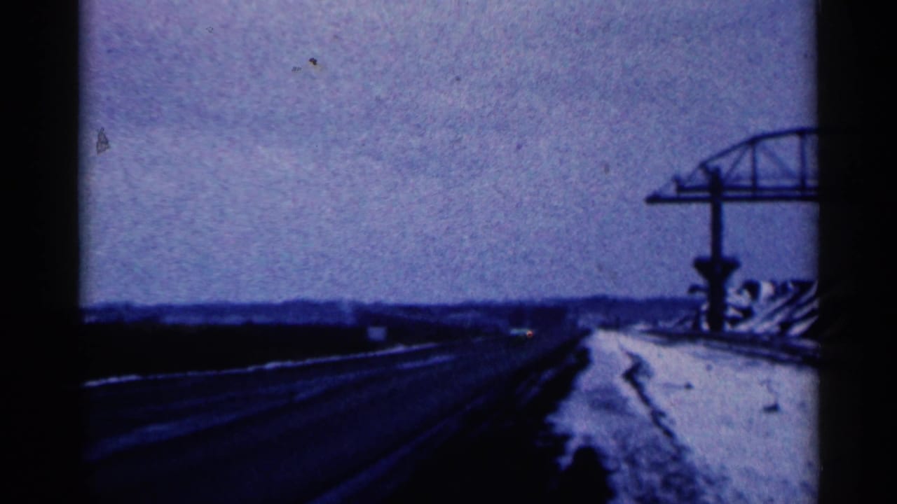 1961 Duluth-area home-movie frame showing a dark winter road, snowbank, and steel roadside framework under cloudy sky.