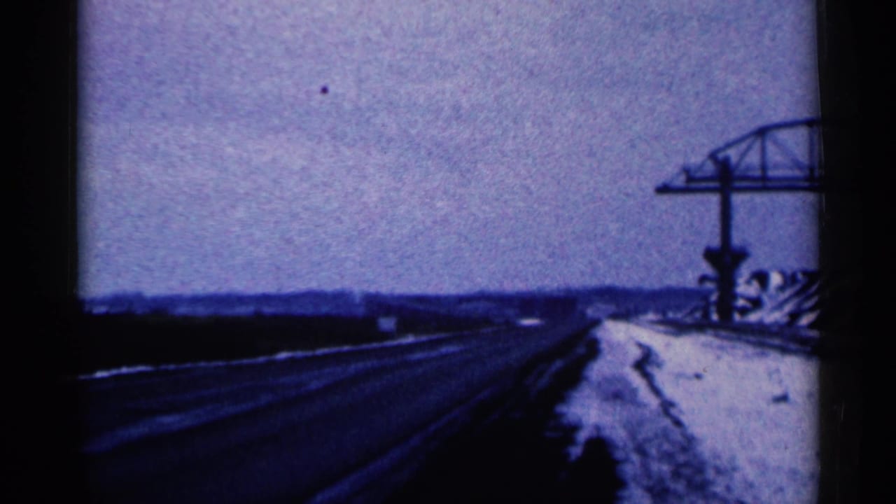 The moving 8mm view looks along a snowy Duluth-area road with dark pavement, low horizon, and roadside steel construction.