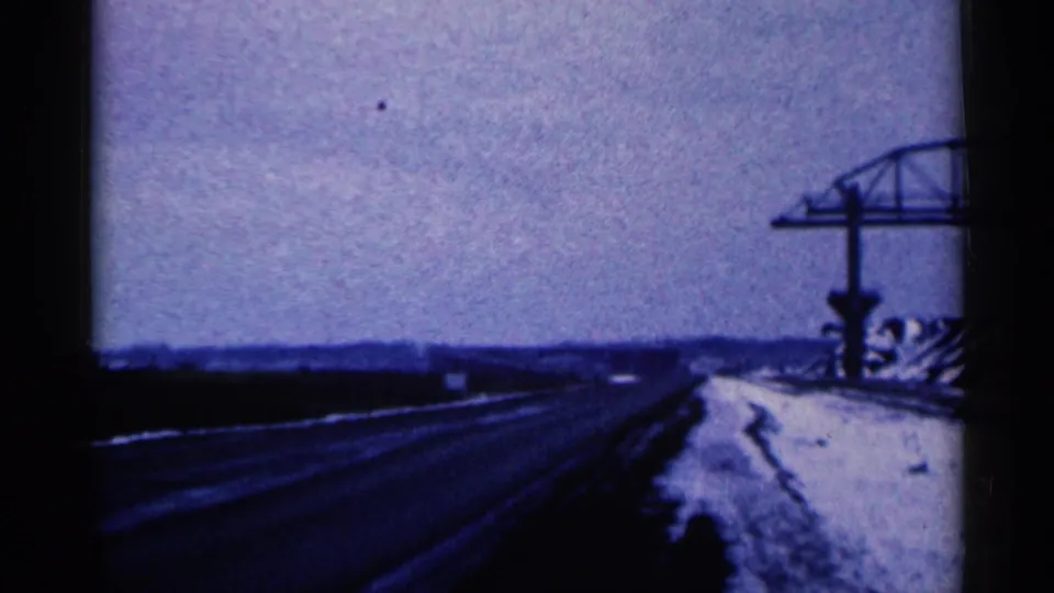 The moving 8mm view looks along a snowy Duluth-area road with dark pavement, low horizon, and roadside steel construction.