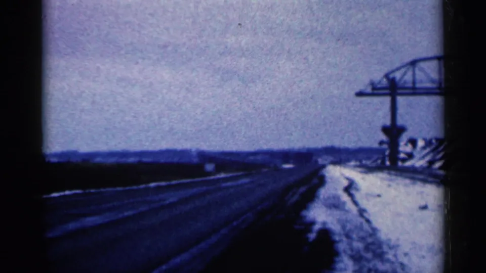 A blue-tinted 1961 home-movie frame shows a snowy road approach with steel construction at the right and a low distant horizon.