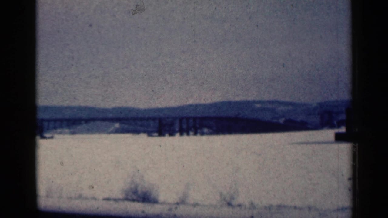 Long bridge crossing a frozen body of water with snowy foreground and distant hills.