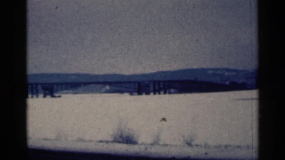 1961 Duluth-area frame with an elevated bridge crossing a frozen expanse, seen through blue-tinted 8mm film grain.