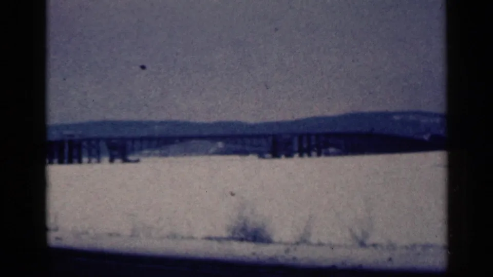 Blue-tinted 1961 Duluth home-movie frame showing a distant bridge, frozen water, snow, and low hills.