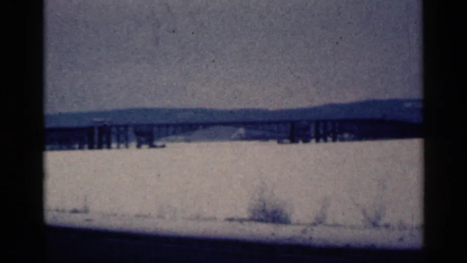 1961 Duluth, Minnesota frame showing bridge supports over frozen water, with snow-covered ground and brush in the foreground.