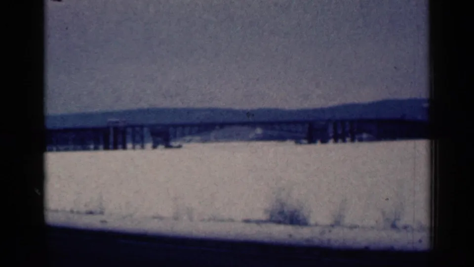 1961 Duluth-area 8mm frame showing snow, a long low bridge or trestle, distant ridge, cloudy sky, and dark film borders.