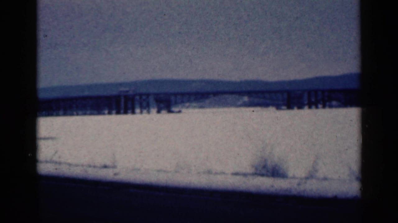 Long bridge spans a snowy or frozen area beneath a cloudy sky and distant ridge.