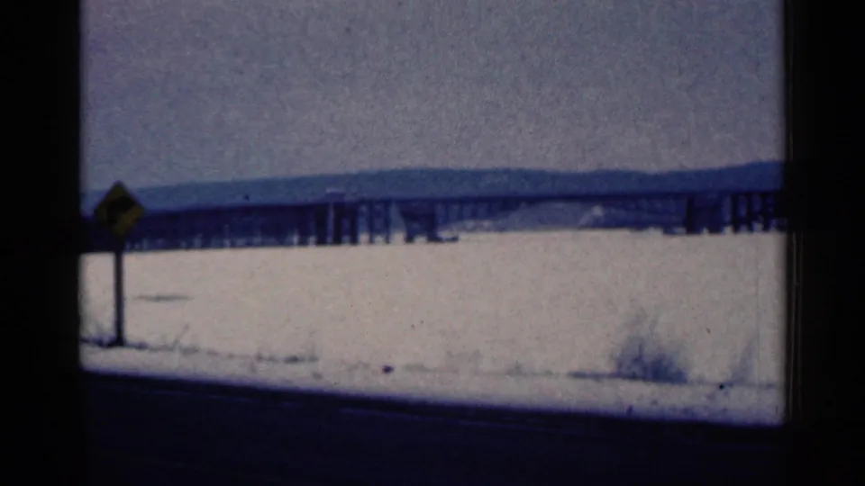 1961 Duluth-area roadside frame with a yellow sign, snowbank, dark road edge, long bridge, ridge line, and winter sky.