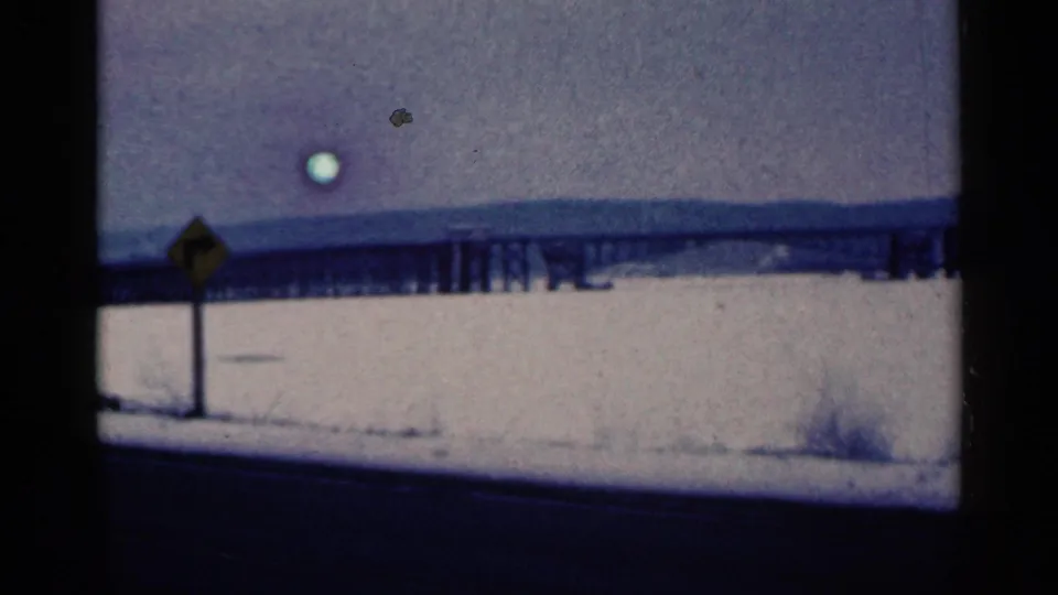 Duluth-area 8mm frame showing a yellow road sign, snow-covered foreground, long bridge or trestle, distant ridge, and visible film artifacts.