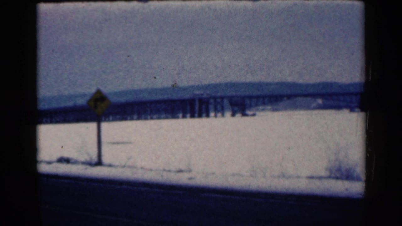 Snowy roadside with a yellow diamond sign and a long bridge in the distance.