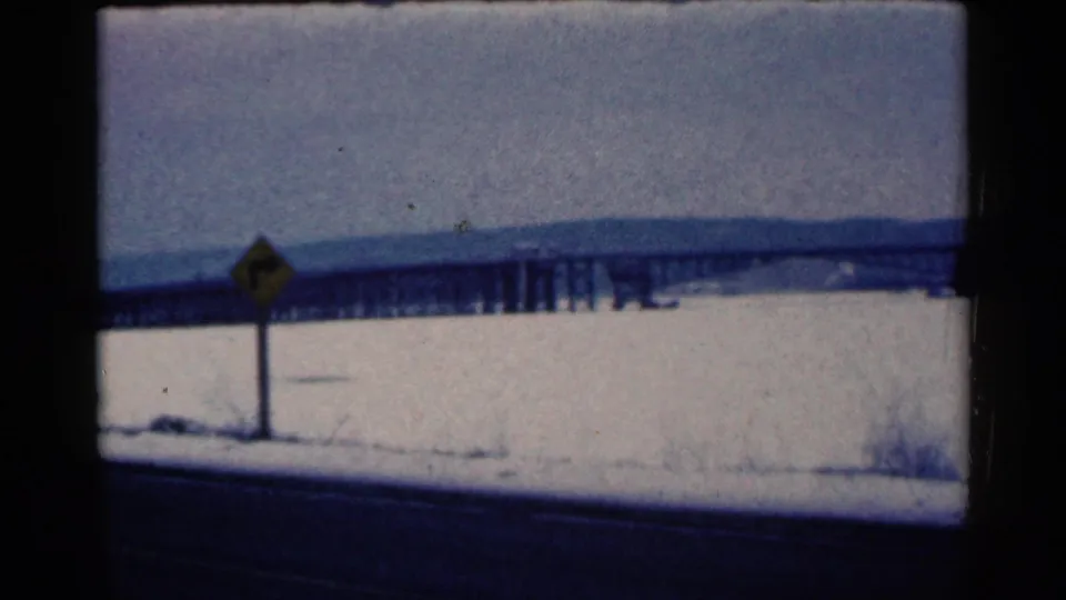 1961 Duluth home-movie frame showing a snow-covered roadside, yellow road sign, dark highway edge, and distant bridge across a frozen expanse.