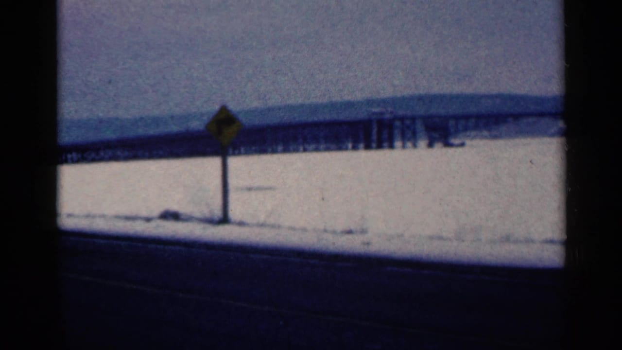 Yellow road sign beside a snowy highway with a bridge stretching across the background.