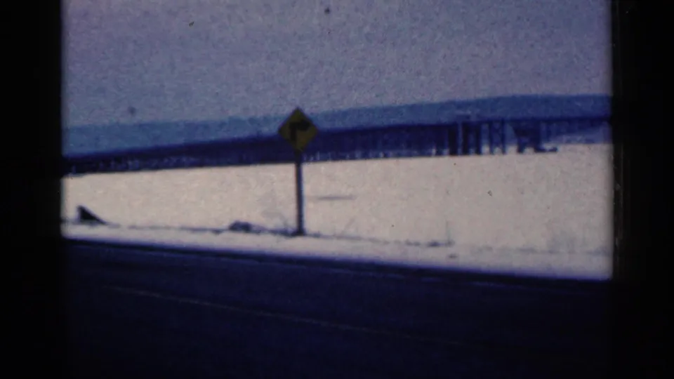 1961 Duluth frame with a yellow roadside sign centered near a snow-covered shoulder, a dark road in front, and a long bridge in the distance.