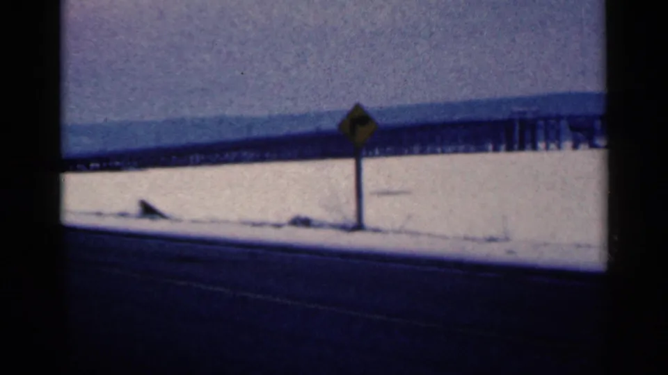 Final 1961 Duluth frame showing the highway foreground, snow-covered ground, yellow signpost, and long bridge receding across the background.
