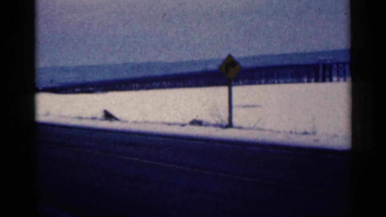 Snowy roadside with a yellow warning sign and distant elevated structure.