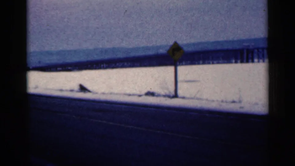 1961 Duluth, Minnesota home-movie frame showing a snow-covered roadside, yellow warning sign, and distant bridge-like structure.
