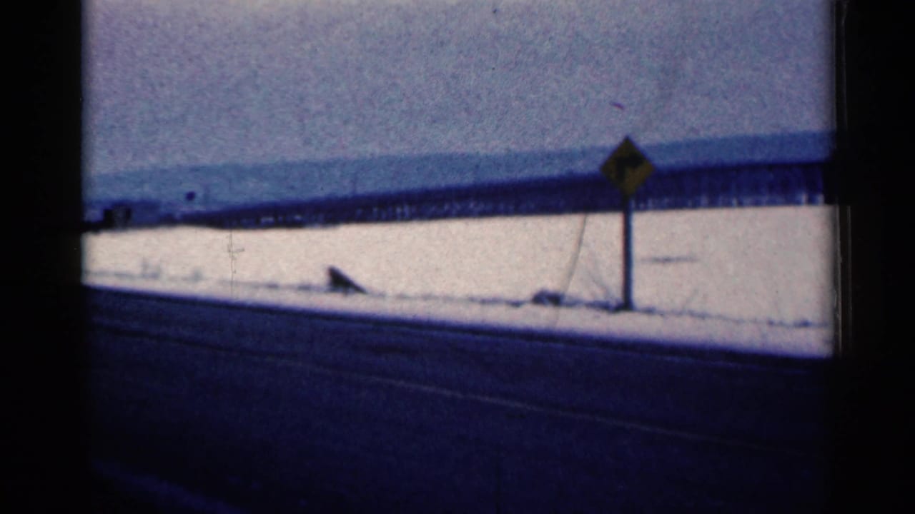 Snowy roadside scene with warning sign, dark road, and distant bridge-like structure.