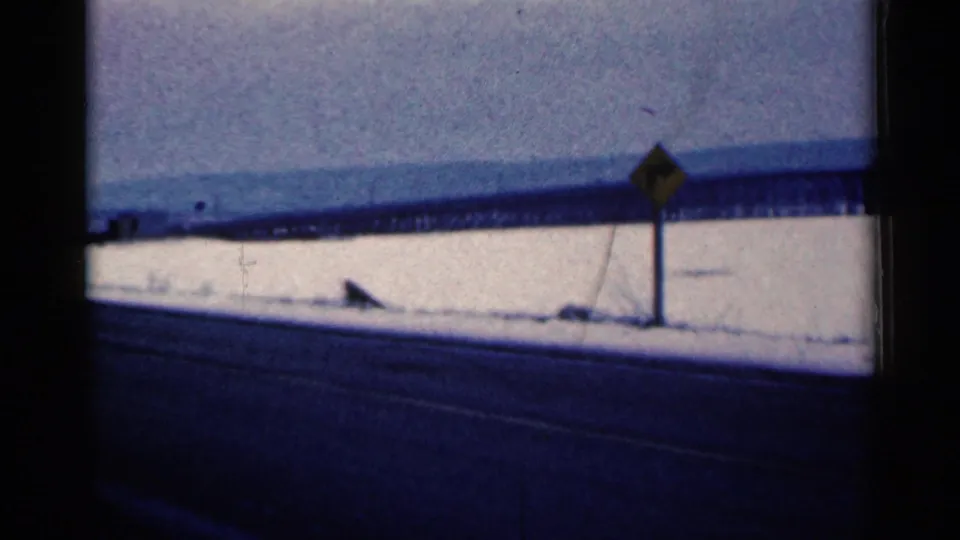 1961 Duluth, Minnesota frame from a moving vehicle showing snowbanks, a roadside warning sign, and distant industrial infrastructure.