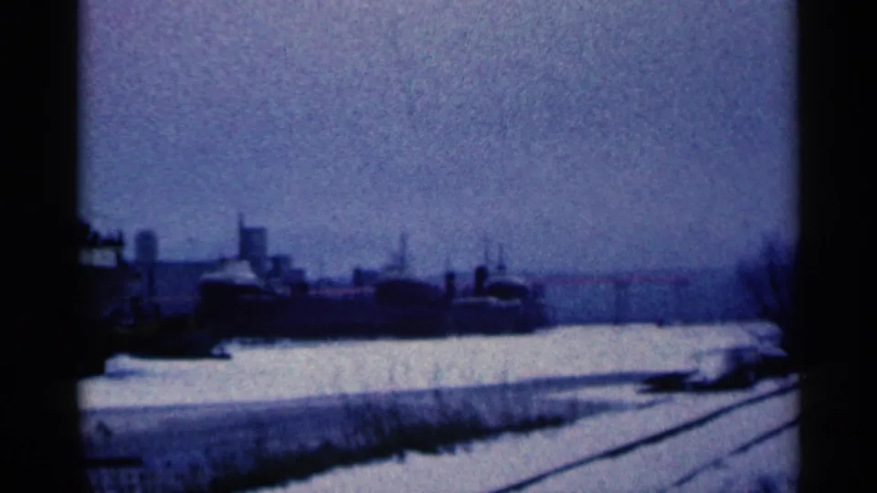 1961 Duluth, Minnesota home-movie frame showing rail tracks beside a snowy industrial waterfront and a large dark vessel or structure.