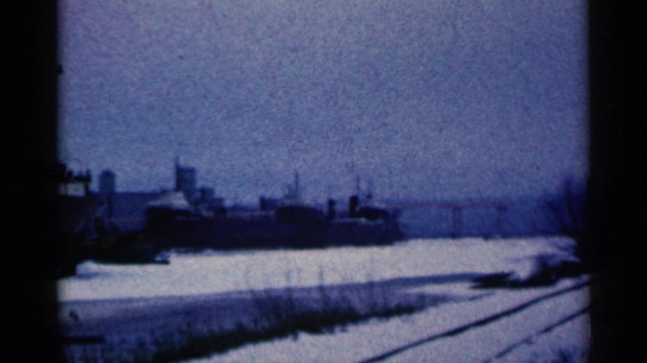 Snowy rail tracks near a distant industrial complex under a gray sky.
