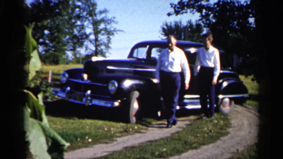 Two adult men stand beside a black sedan on a curved grassy drive in 1961 Duluth home-movie footage.
