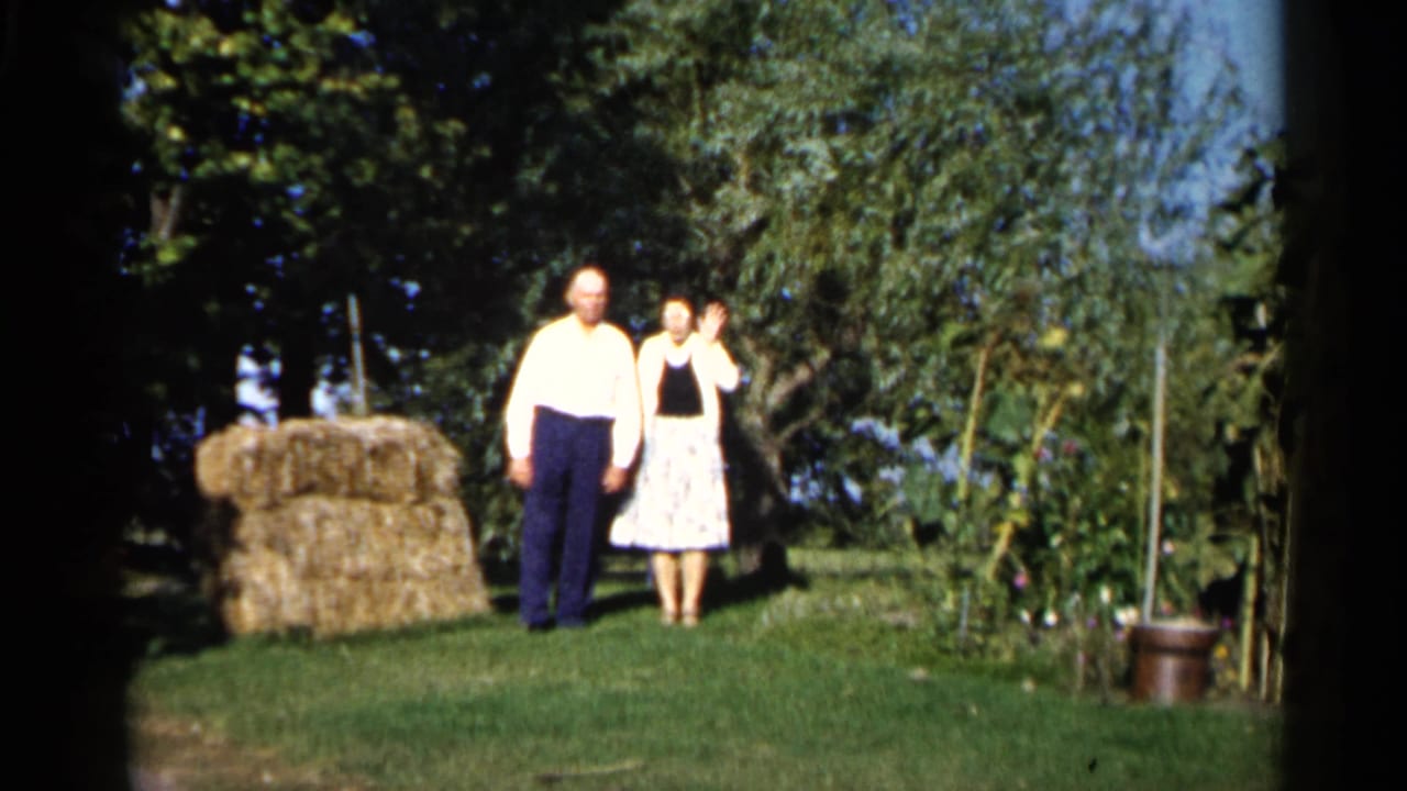 In 1961 Duluth home-movie footage, an older couple stands in a sunny backyard near a hay bale, garden plants, and trees.