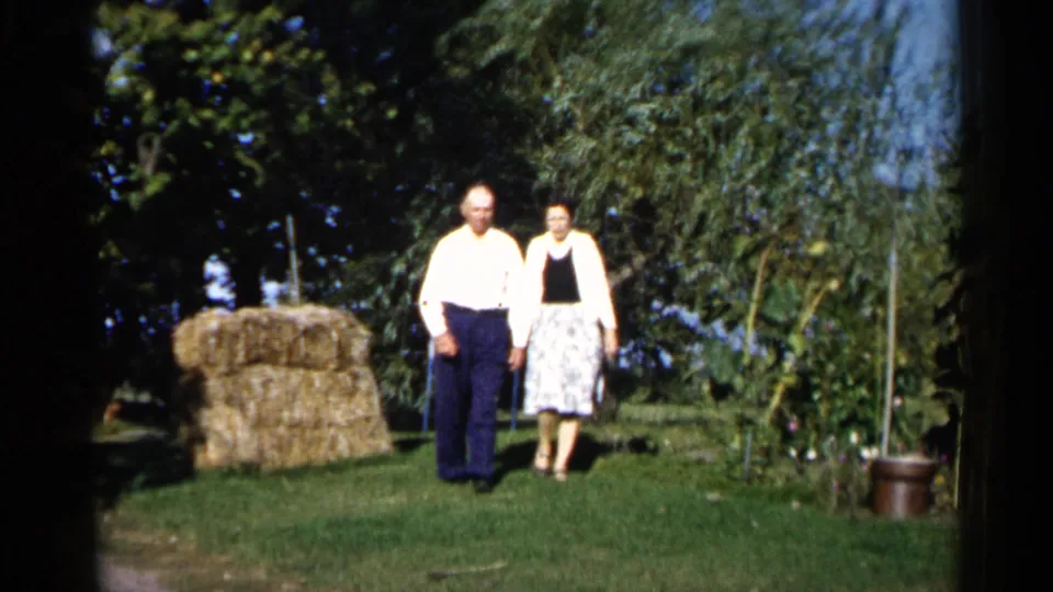 The older couple walks toward the camera across the lawn, with the hay bale to their left and garden plants behind them.
