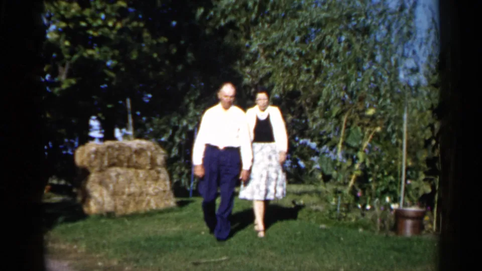 The couple approaches the camera in the 1961 Duluth backyard scene, framed by trees, a hay bale, garden growth, and bright sunlight.