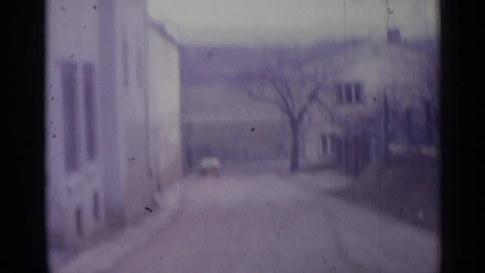 1961 Spanish village exterior with a pale building close to camera, road receding past a bare tree, and low houses in the distance.