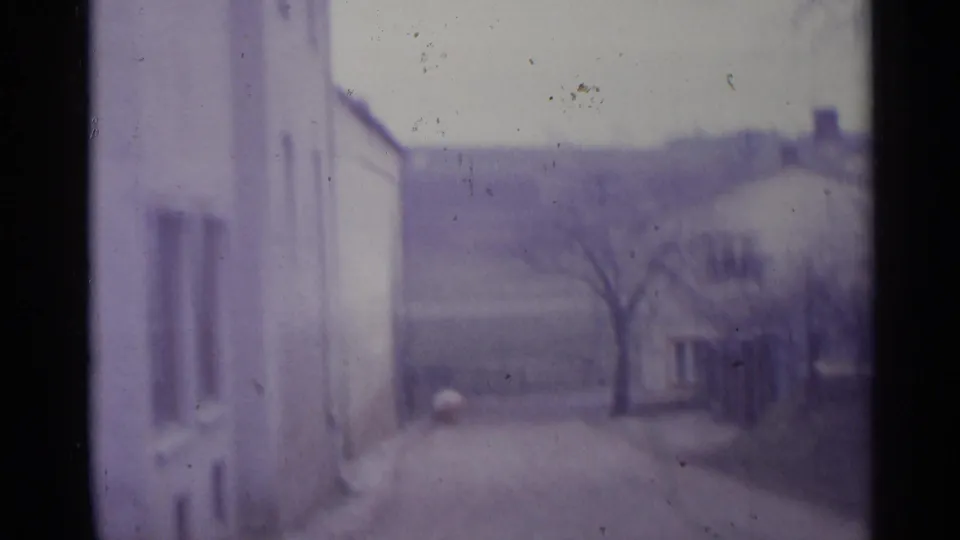 Soft 8mm frame from 1961 Spain showing the road, pale stucco-like walls, a bare roadside tree, and low buildings under gray light.