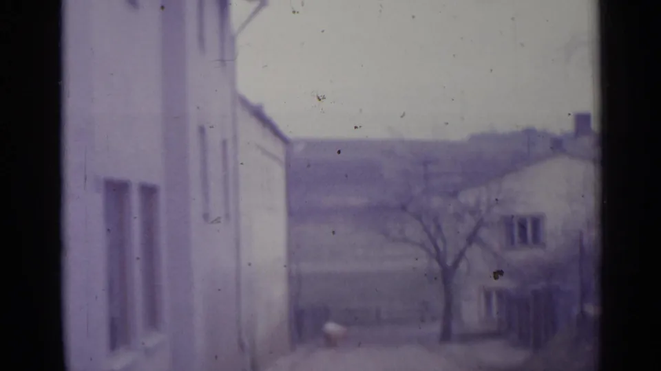 1961 Spain home-movie view with a pale wall in the foreground, unpaved road, bare tree, and distant low buildings in soft focus.