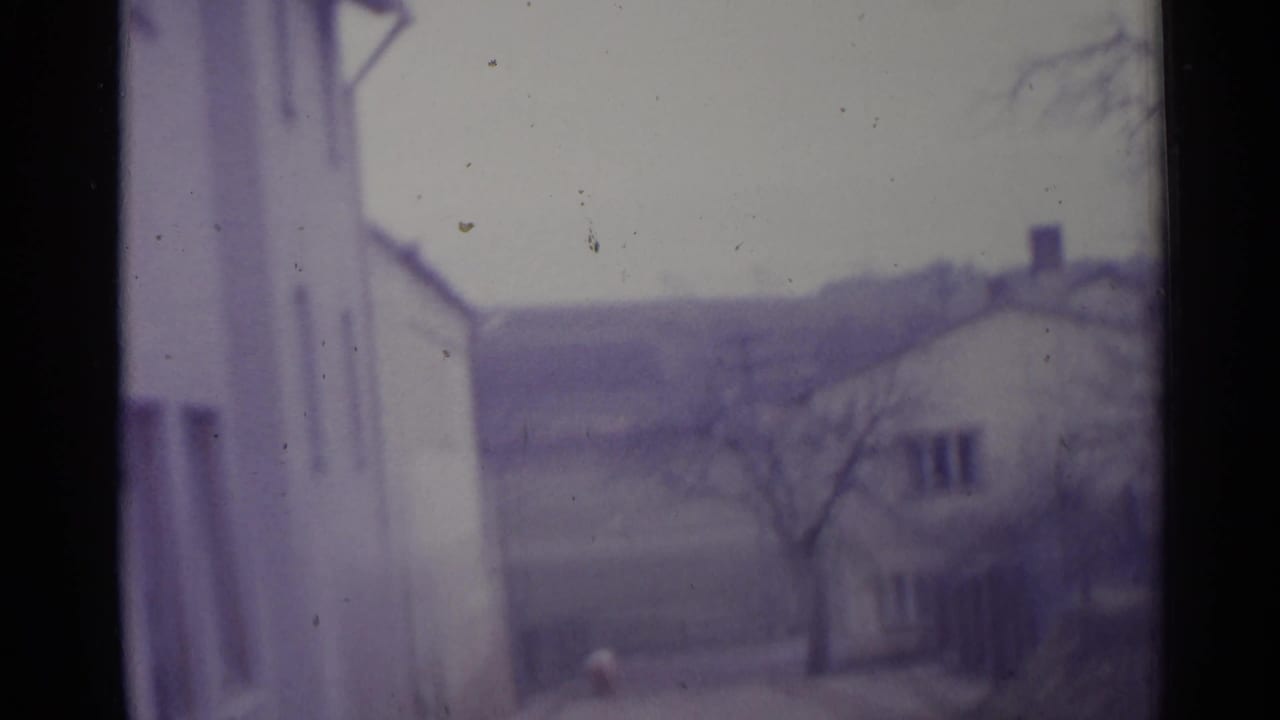 Soft view between white buildings toward bare trees and a distant hillside under cloudy sky.