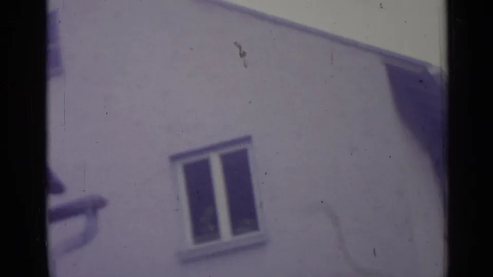 1961 Spain home-movie frame showing a white residential wall, closed window, and sloped roof edge in aged 8mm color.