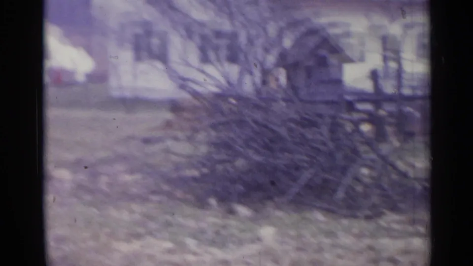A large brush pile rests in a rural yard, with wooden fencing or outbuildings visible in the soft 1961 Spain footage.