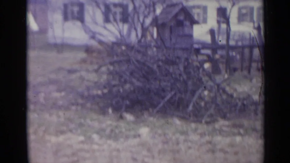 1961 Spain home-movie frame showing cut branches in a yard, a rough fence, bare trees, and a white house with shutters.
