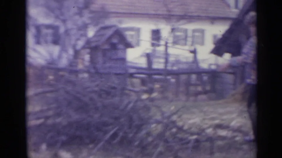 1961 Spain home-movie view of a branch pile beside a rough fence, small wooden structure, and white house with shutters.