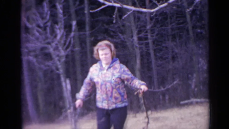 1961 Spain home-movie frame of an adult woman in a colorful patterned jacket walking through bare woods while holding branches or sticks.