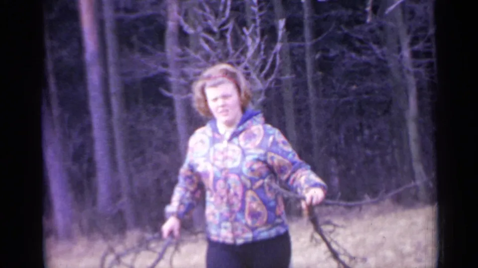 1961 Spain home-movie frame showing an adult woman facing the camera in a patterned jacket, holding a long branch in a leafless forest.