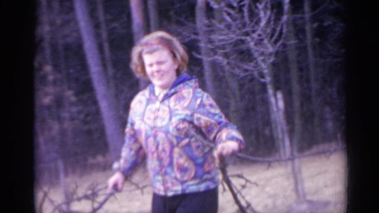 Woman in a colorful jacket carries branches in a wooded area.