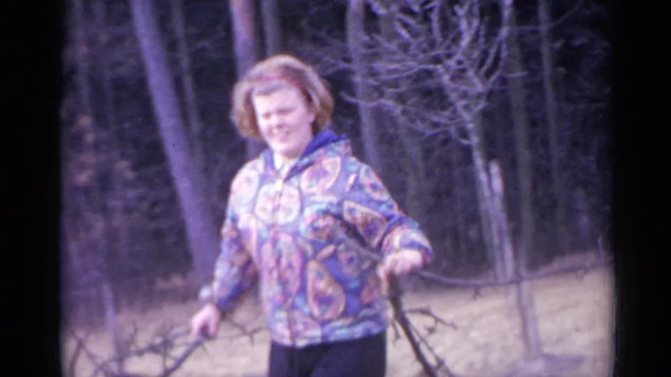 In 1961 Spain home-movie footage, a woman in a colorful patterned jacket carries fallen branches among bare trees.