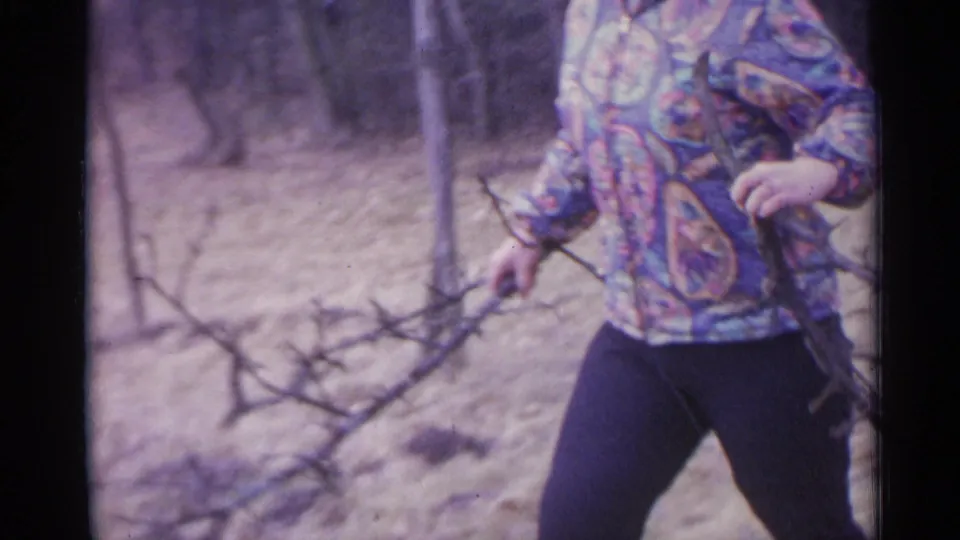 A cropped 8mm frame shows the woman holding a bundle of branches, with dry ground and trees behind her.
