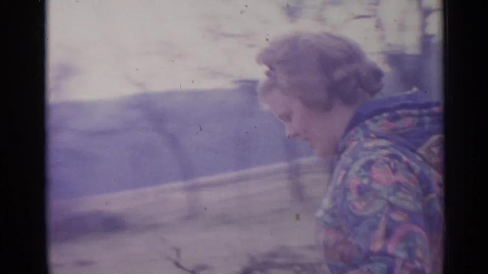 The woman leans downward with branches in hand, seen in soft 8mm focus against an outdoor wooded background.
