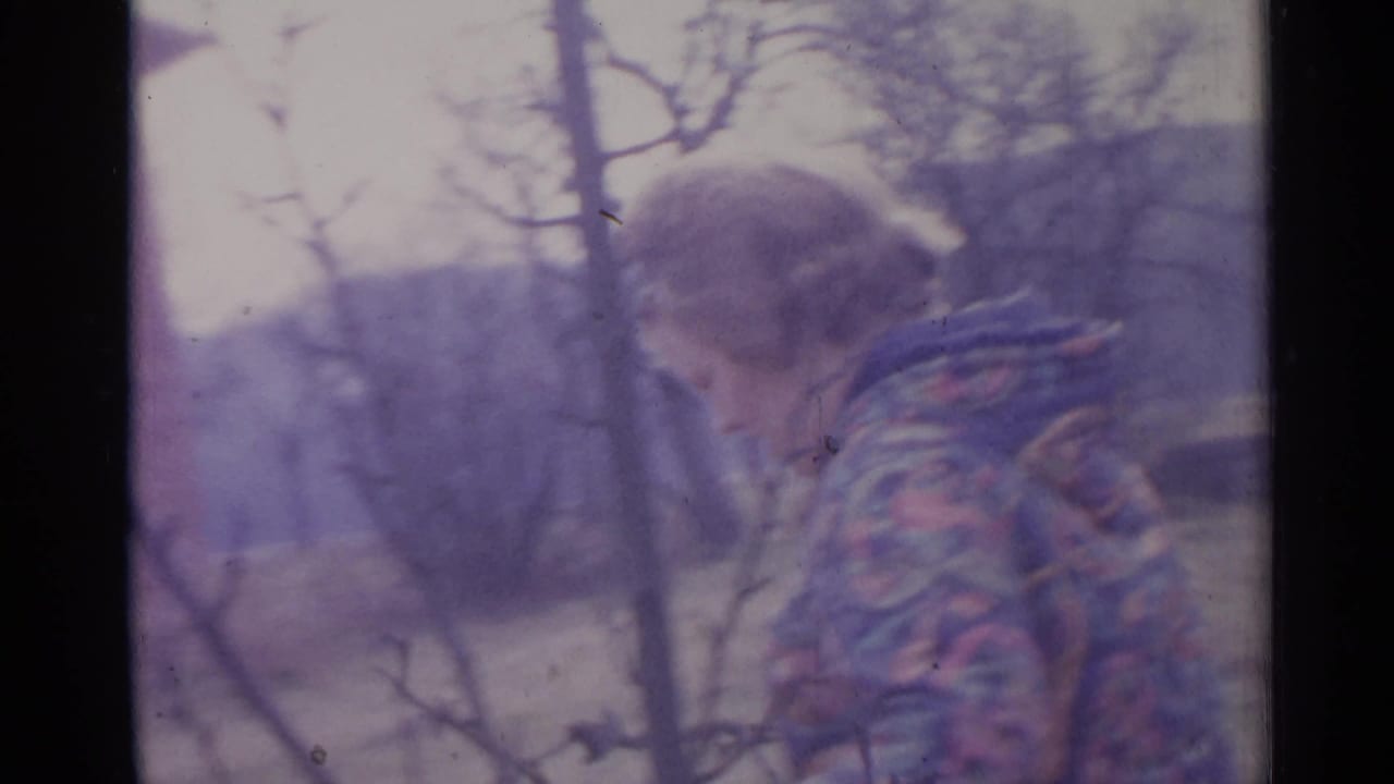Person in a patterned jacket seen near bare branches in a rural yard.