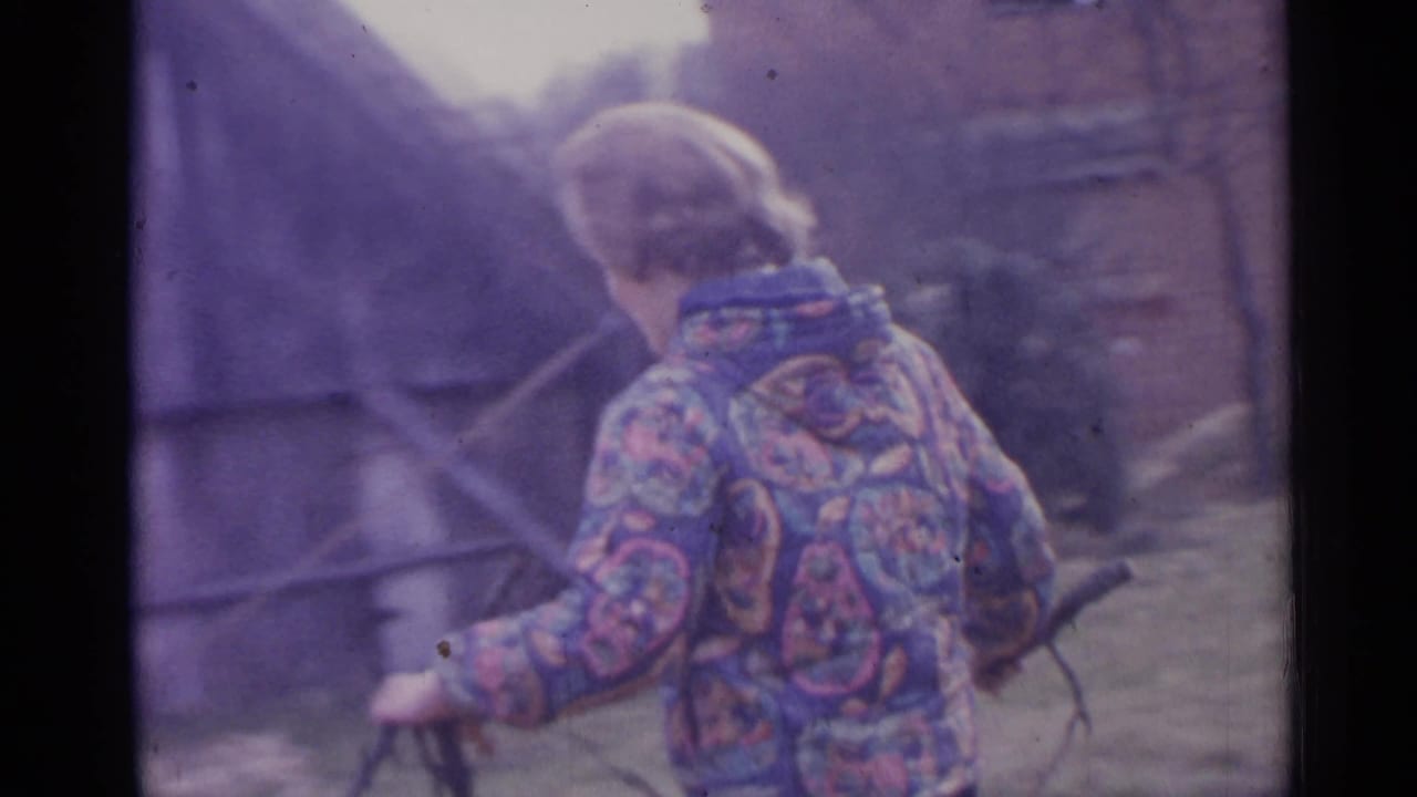 Person in a patterned jacket walks beside rustic buildings while holding a branch or tool.