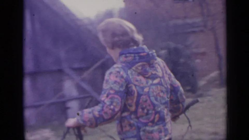 A person in a patterned jacket walks through a Spanish farmstead area, holding a branch or long tool near rustic buildings.