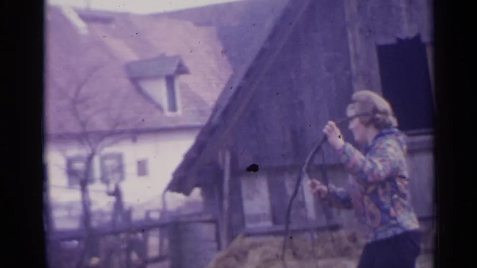The person raises a branch or tool beside a rustic outbuilding, with a tiled-roof house visible in the background.