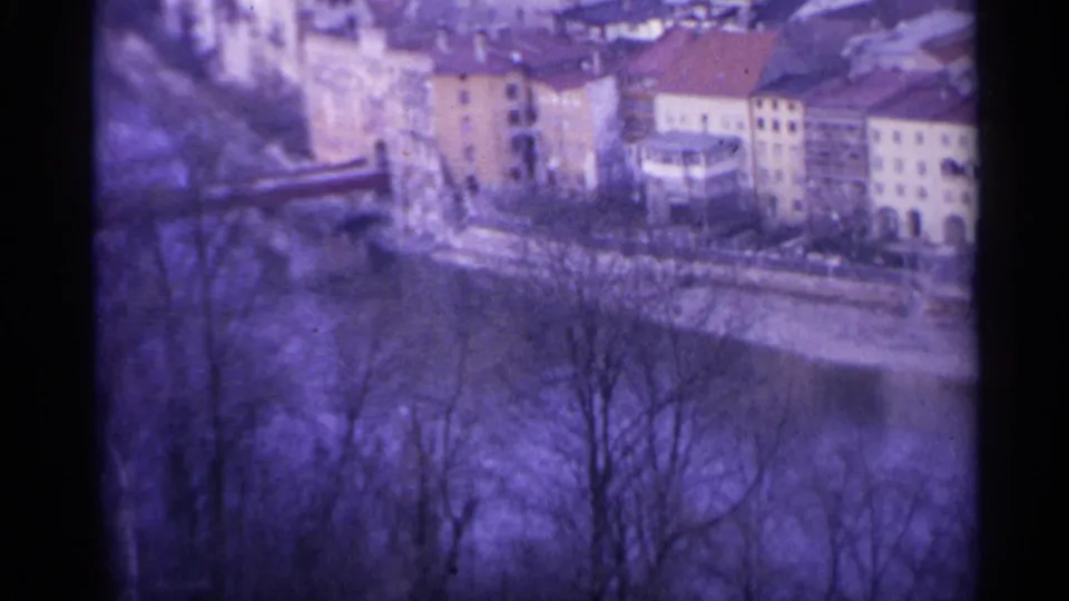1961 Spain 8mm frame showing colorful riverbank buildings and red roofs beyond leafless foreground trees.