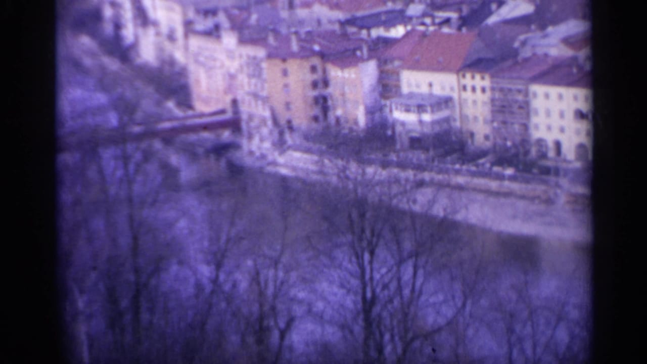 1961 8mm view in Spain of a river, small bridge, pale town buildings, red-tiled roofs, and leafless foreground trees.